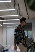 A person with tattoos and wearing a colorful floral shirt is standing in a modern room with industrial-style ceilings and hanging fluorescent lights. The room has a casual atmosphere, with visible objects like a chair and some green plants. Light streams through a window, adding to the ambient lighting.