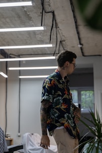A person with tattoos and wearing a colorful floral shirt is standing in a modern room with industrial-style ceilings and hanging fluorescent lights. The room has a casual atmosphere, with visible objects like a chair and some green plants. Light streams through a window, adding to the ambient lighting.