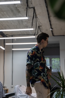 A person with tattoos and wearing a colorful floral shirt is standing in a modern room with industrial-style ceilings and hanging fluorescent lights. The room has a casual atmosphere, with visible objects like a chair and some green plants. Light streams through a window, adding to the ambient lighting.