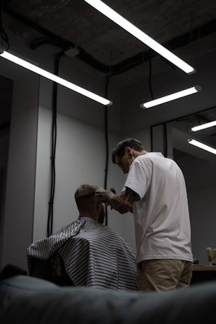 A barber, wearing glasses and a white T-shirt, is giving a haircut to a client seated in a chair. The client is covered with a striped cape. The setting is a modern and dimly lit barbershop with exposed ceilings and fluorescent lights. Mirrors and workstations with barber tools can be seen in the background.