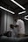 A barber, wearing glasses and a white T-shirt, is giving a haircut to a client seated in a chair. The client is covered with a striped cape. The setting is a modern and dimly lit barbershop with exposed ceilings and fluorescent lights. Mirrors and workstations with barber tools can be seen in the background.