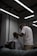 A barber, wearing glasses and a white T-shirt, is giving a haircut to a client seated in a chair. The client is covered with a striped cape. The setting is a modern and dimly lit barbershop with exposed ceilings and fluorescent lights. Mirrors and workstations with barber tools can be seen in the background.