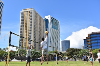 A group of people are playing volleyball outdoors on a grassy field with a volleyball net set up. Tall modern buildings and skyscrapers form the background, under a clear blue sky. Lush green trees surround the area, creating a bright and lively atmosphere.