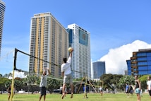 A group of people are playing volleyball outdoors on a grassy field with a volleyball net set up. Tall modern buildings and skyscrapers form the background, under a clear blue sky. Lush green trees surround the area, creating a bright and lively atmosphere.