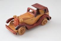 A handcrafted wooden toy car featuring a classic design with exposed fenders, open-top, and a retro style. The wood grain is visible, alternating between light and dark tones, with intricate details on headlights and wheels.