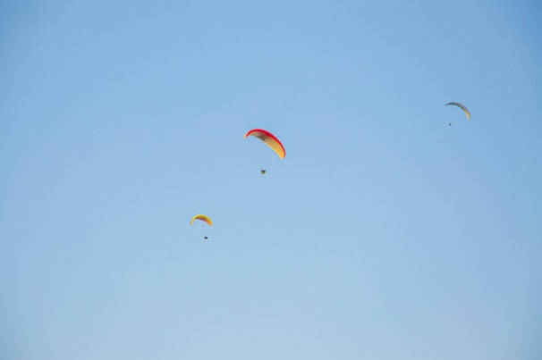 Three paragliders are soaring high against a clear blue sky. Each paraglider features vibrant colors, with the largest one having red and yellow accents. The smaller two also display bright colors as they float gracefully in the open air.