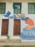 A mural painted on a building wall featuring a person in an orange shirt kneeling by a representation of water, surrounded by blue leaves and flowers. The art also depicts a blue bird in flight. Two brown wooden doors are beneath two green shuttered windows. A small potted plant is placed in front of one door.