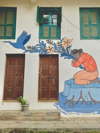 A mural painted on a building wall featuring a person in an orange shirt kneeling by a representation of water, surrounded by blue leaves and flowers. The art also depicts a blue bird in flight. Two brown wooden doors are beneath two green shuttered windows. A small potted plant is placed in front of one door.