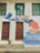 A mural painted on a building wall featuring a person in an orange shirt kneeling by a representation of water, surrounded by blue leaves and flowers. The art also depicts a blue bird in flight. Two brown wooden doors are beneath two green shuttered windows. A small potted plant is placed in front of one door.