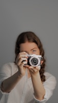 A person with long hair poses while holding a digital camera up to their face, creating a focused and engaging image. The background appears to be a plain, neutral color, and the lighting is even, highlighting the person and the camera.