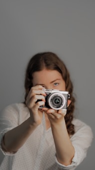 A person with long hair poses while holding a digital camera up to their face, creating a focused and engaging image. The background appears to be a plain, neutral color, and the lighting is even, highlighting the person and the camera.