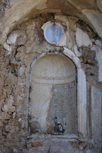 An old, weathered stone niche in a crumbling wall with a small circular decorative element above. The niche is empty except for some dry vegetation and a painted image of a cat on the inner surface. The structure shows significant signs of deterioration with visible cracks and missing pieces.