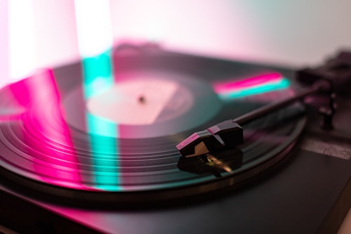 A close-up view of a vinyl record on a turntable with a tonearm and needle. The scene is illuminated with colorful lights, predominantly featuring shades of pink and teal. The focus is on the spinning record and the stylus.