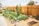 A vibrant community garden is surrounded by a tall wooden fence. Numerous raised wooden garden beds contain lush green plants, while freshly tilled earth is visible in some plots. Several rocks are placed decoratively along the garden's pathway. In the background, there are trees and a few houses, providing a suburban feel to the scene.