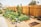A vibrant community garden is surrounded by a tall wooden fence. Numerous raised wooden garden beds contain lush green plants, while freshly tilled earth is visible in some plots. Several rocks are placed decoratively along the garden's pathway. In the background, there are trees and a few houses, providing a suburban feel to the scene.