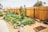 A vibrant community garden is surrounded by a tall wooden fence. Numerous raised wooden garden beds contain lush green plants, while freshly tilled earth is visible in some plots. Several rocks are placed decoratively along the garden's pathway. In the background, there are trees and a few houses, providing a suburban feel to the scene.