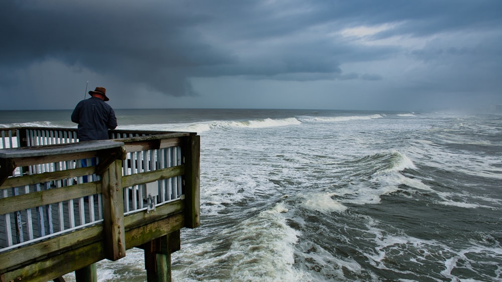 person in black jacket sitting on brown wooden dock over sea waves during daytime