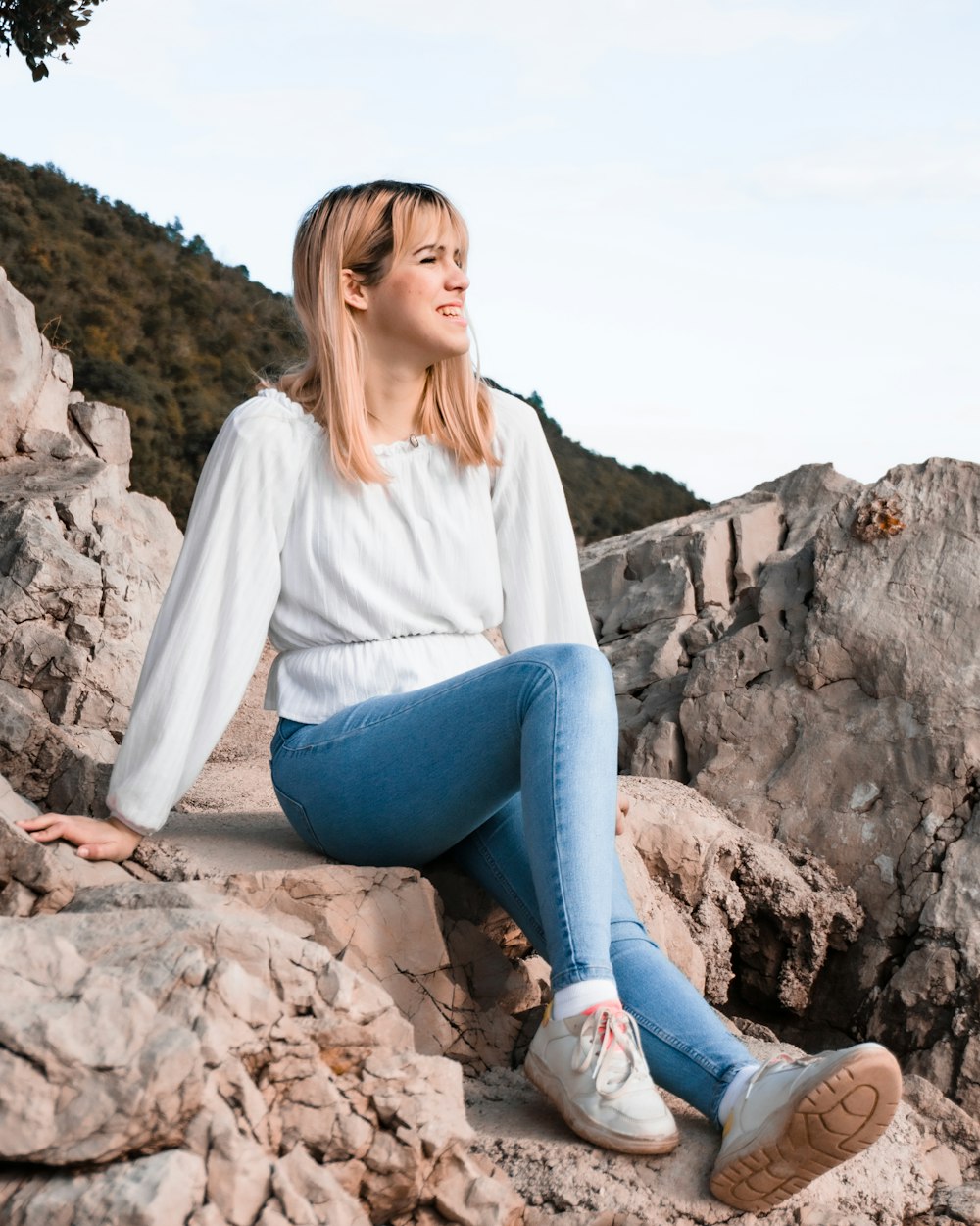 woman in white long sleeve shirt and blue denim jeans sitting on rock during daytime