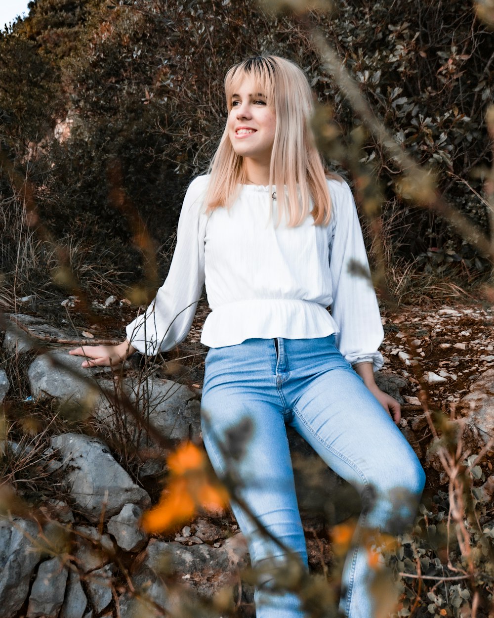 woman in white long sleeve shirt and blue denim jeans sitting on brown dried leaves