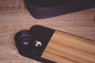 A smartphone case with a lens attachment is set on a textured wooden surface. The case features a wooden panel design and a camera lens module protruding from its back. A fabric-covered object, possibly another case, is partially visible in the upper section.