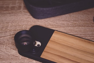 A smartphone case with a lens attachment is set on a textured wooden surface. The case features a wooden panel design and a camera lens module protruding from its back. A fabric-covered object, possibly another case, is partially visible in the upper section.