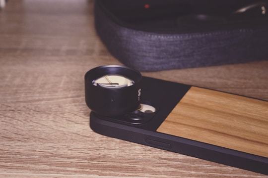 A smartphone with a wooden back panel is positioned on a wooden surface. Attached to the phone is a large black external lens, suggesting it is set up for photography. In the background, there is a soft-focus image of a fabric pouch, likely used for storing the lens.