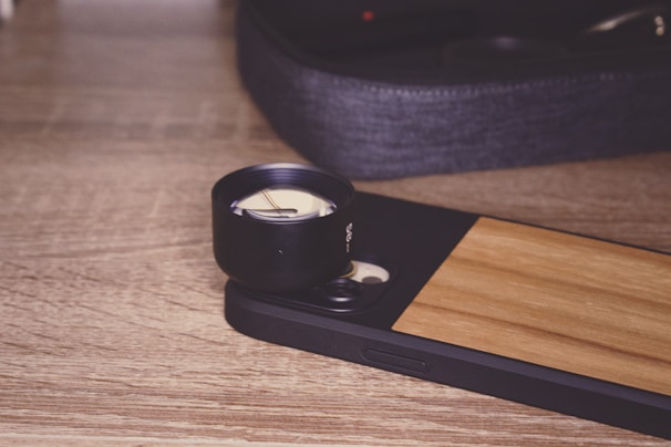 A smartphone with a wooden back panel is positioned on a wooden surface. Attached to the phone is a large black external lens, suggesting it is set up for photography. In the background, there is a soft-focus image of a fabric pouch, likely used for storing the lens.