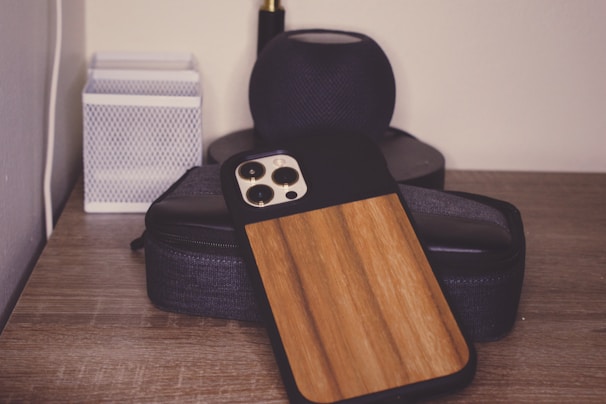 A wooden phone case is resting on a stack of black electronic devices on a wooden surface. There is a white rectangular item with a textured design situated on the left.