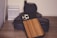 A wooden phone case is resting on a stack of black electronic devices on a wooden surface. There is a white rectangular item with a textured design situated on the left.