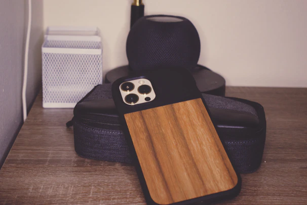 A wooden phone case is resting on a stack of black electronic devices on a wooden surface. There is a white rectangular item with a textured design situated on the left.
