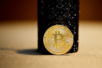 A gold-colored Bitcoin coin leaning against a dark textured cylindrical object. The focus is primarily on the Bitcoin symbol in the center of the coin, with the background slightly blurred, emphasizing the coin's details.