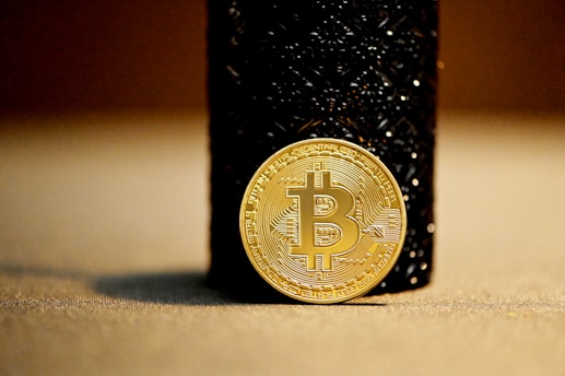 A gold-colored Bitcoin coin leaning against a dark textured cylindrical object. The focus is primarily on the Bitcoin symbol in the center of the coin, with the background slightly blurred, emphasizing the coin's details.