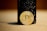 A gold-colored Bitcoin coin leaning against a dark textured cylindrical object. The focus is primarily on the Bitcoin symbol in the center of the coin, with the background slightly blurred, emphasizing the coin's details.