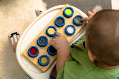 A child wearing a green shirt is sitting at a table, interacting with a tactile toy. The toy consists of a wooden board with several blue circular frames containing different textured materials like fabric and sandpaper. The child is focused on touching these textures.
