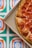 A pizza with pepperoni topping inside a pizza box. The background is a vivid, geometric pattern with green, blue, and orange shapes.