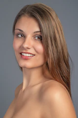 A woman with long brown hair smiles against a neutral gray background. Her skin appears smooth and she has light makeup on, emphasizing her features.