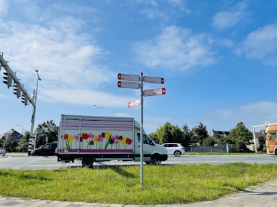 A street scene featuring a colorful truck with floral patterns parked on the roadside. There are multiple road signs pointing towards different directions and indicating destinations such as Maasluis and Rotterdam. The sky is blue with some scattered clouds, and the atmosphere is calm with greenery surrounding the area.