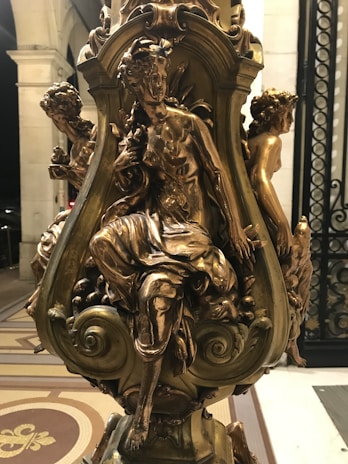 A beautifully detailed bronze sculpture featuring three elegantly posed figures in a classical style. Each figure is finely sculpted with intricate detailing on their clothing and expressions. The sculpture is mounted on a decorative base with ornamental patterns, set against a backdrop of architectural columns and patterned flooring.