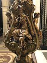 A beautifully detailed bronze sculpture featuring three elegantly posed figures in a classical style. Each figure is finely sculpted with intricate detailing on their clothing and expressions. The sculpture is mounted on a decorative base with ornamental patterns, set against a backdrop of architectural columns and patterned flooring.