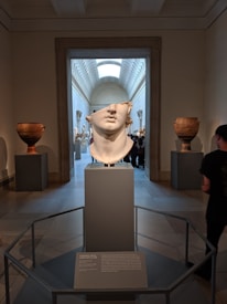 A sculpture of a fragmented classical head displayed on a pedestal in a museum gallery. The room features large pottery pieces on either side and an ornate archway leading to a hall filled with more classical sculptures. The gallery is lit by overhead skylights.