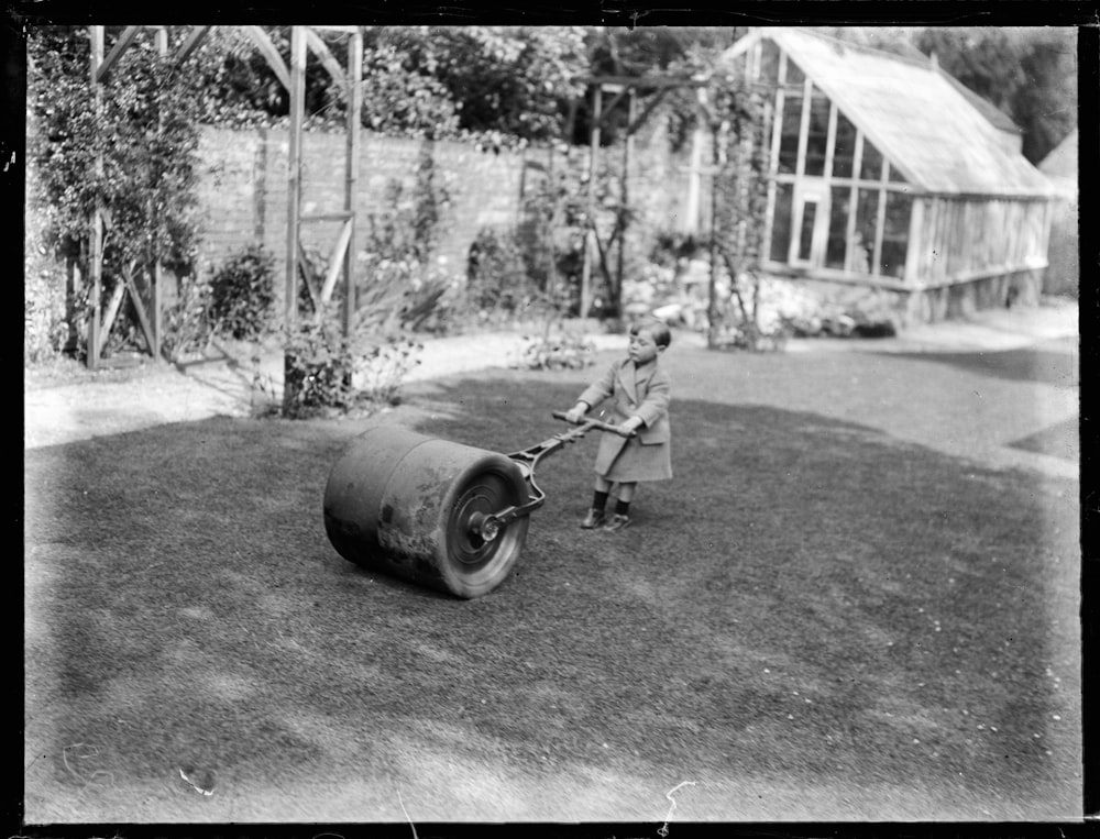 grayscale photo of man in black jacket and pants standing beside black metal wheel barrow