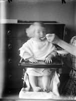 A young child sitting in a high chair is being fed by an adult. The child's face is slightly blurred, suggesting movement as they eat. The adult's hand is holding a spoon near the child's mouth, and they are wearing a plaid shirt, partially visible on the right. The background features a dark wooden dresser.