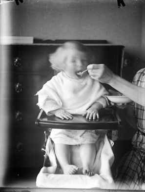 A young child sitting in a high chair is being fed by an adult. The child's face is slightly blurred, suggesting movement as they eat. The adult's hand is holding a spoon near the child's mouth, and they are wearing a plaid shirt, partially visible on the right. The background features a dark wooden dresser.