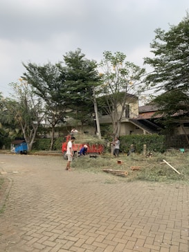 A few people are working on cutting and clearing branches from trees. A red truck is parked nearby, loaded with chopped branches. The scene takes place on a paved road with trees and a house in the background. The sky is overcast, adding a neutral tone to the environment.