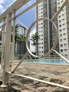 A swimming pool area surrounded by tall residential buildings. The pool is enclosed by a white metal fence, featuring geometric patterns. Palm trees add a tropical touch to the urban environment. The sky is clear with a few clouds.