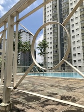 A swimming pool area surrounded by tall residential buildings. The pool is enclosed by a white metal fence, featuring geometric patterns. Palm trees add a tropical touch to the urban environment. The sky is clear with a few clouds.
