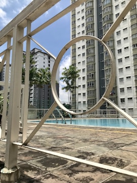 A swimming pool area surrounded by tall residential buildings. The pool is enclosed by a white metal fence, featuring geometric patterns. Palm trees add a tropical touch to the urban environment. The sky is clear with a few clouds.