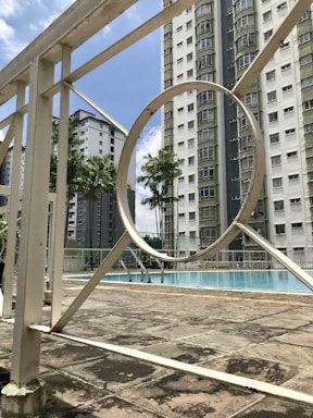 A swimming pool area surrounded by tall residential buildings. The pool is enclosed by a white metal fence, featuring geometric patterns. Palm trees add a tropical touch to the urban environment. The sky is clear with a few clouds.