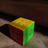 A cube puzzle with three visible colored faces is placed on a marbled surface. The colors on the cube are green, yellow, and orange, catching light from one side and casting a shadow on the other side.