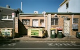 An urban alleyway featuring a small brick building with graffiti on the walls. Several recycling and trash bins are lined up against the building, with colorful graffiti marking the exterior. The area is set under a clear blue sky, creating a stark contrast between the gritty urban feel and the natural backdrop.